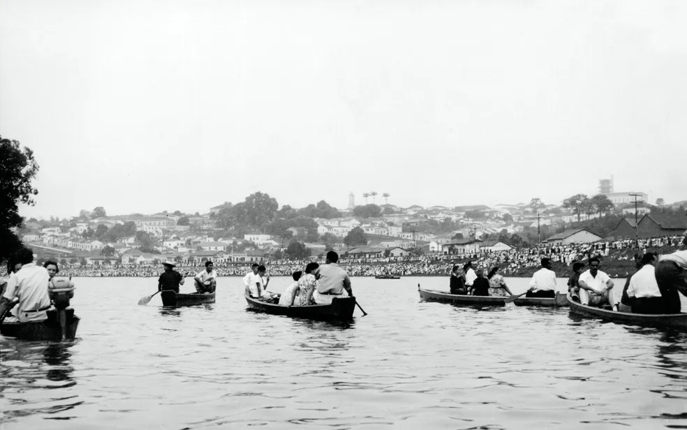 A imagem em preto e branco mostra uma cena movimentada em um lago ou rio, com várias canoas ocupadas por pessoas. Ao fundo, uma cidade se estende sobre uma colina, com casas, árvores e alguns prédios mais altos visíveis. A paisagem é calma e o céu está claro. As pessoas nas canoas parecem estar desfrutando de um passeio ou evento recreativo, com algumas delas remando e outras conversando. A vestimenta das pessoas sugere uma época passada, possivelmente meados do século XX. O contraste entre a água calma, as pessoas nas canoas e a cidade ao fundo cria uma composição visual interessante. A atmosfera geral da imagem é tranquila e nostálgica.