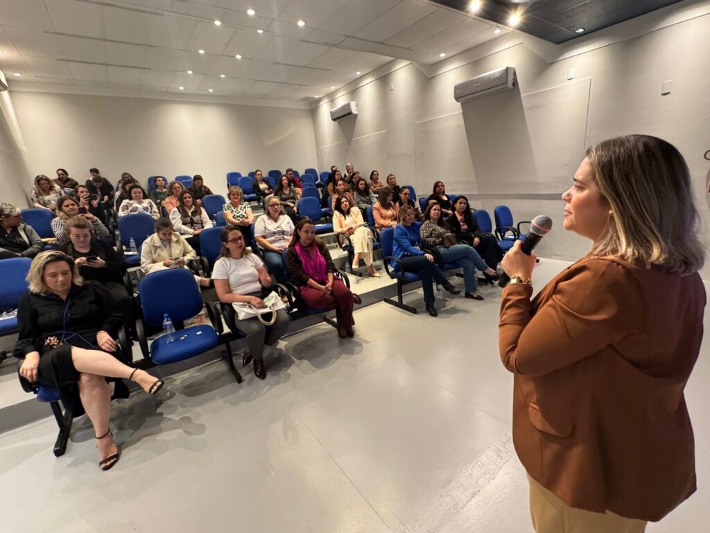 A imagem mostra uma sala de conferências com uma plateia de mulheres sentadas em cadeiras azuis, voltadas para uma mulher que está em pé, falando em um microfone. A mulher que fala usa um blazer marrom e calças bege, e tem o cabelo loiro na altura dos ombros. A plateia é composta por mulheres de diversas idades, a maioria vestindo roupas casuais. As cadeiras estão dispostas em fileiras em degraus, e a sala parece ter iluminação artificial. Há dois aparelhos de ar condicionado nas paredes.