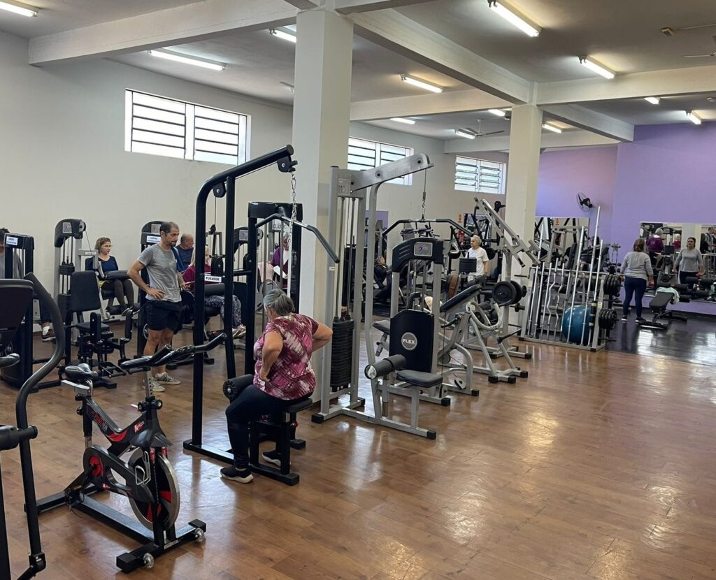 A imagem mostra uma academia de ginástica relativamente espaçosa com uma variedade de equipamentos de exercícios. O chão é de madeira escura e polida, e as paredes são pintadas de um tom lavanda claro. O teto apresenta luminárias fluorescentes, fornecendo iluminação abundante ao espaço. Vários indivíduos estão presentes na academia, a maioria aparentemente adultos mais velhos, engajados em diferentes atividades de exercícios. Algumas pessoas estão usando máquinas de musculação, enquanto outras parecem estar descansando ou conversando. Há uma mistura de equipamentos de musculação, incluindo máquinas de peso, aparelhos de musculação e bicicletas ergométricas. O ambiente parece limpo e bem organizado, com os equipamentos dispostos de forma lógica e espaçosa. Há um grande espelho na parede do fundo, refletindo parte da academia e algumas pessoas. No geral, a imagem transmite uma sensação de calma e atividade física moderada, sugerindo uma academia voltada para um público mais maduro ou com foco em exercícios de baixo impacto.