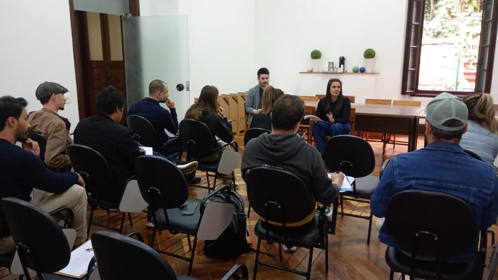 A imagem mostra uma sala de aula ou sala de reuniões com várias pessoas sentadas em cadeiras em círculo. Há uma mesa na parte de trás da sala, e uma janela com vista para o exterior. As pessoas parecem estar envolvidas numa discussão ou apresentação. A sala é bem iluminada e organizada. As pessoas estão vestidas de forma casual, e o ambiente parece tranquilo e profissional. Há um homem e uma mulher sentados na frente do grupo, possivelmente líderes ou apresentadores. O espaço parece ser um local de aprendizagem ou trabalho colaborativo.
