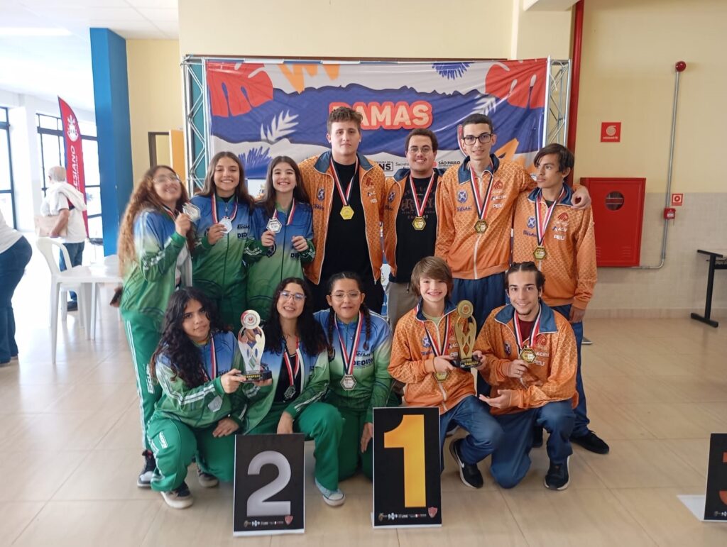 A imagem mostra um grupo de jovens, possivelmente estudantes, posando para uma foto após uma competição. Eles estão vestidos com uniformes de equipe, alguns em tons de verde e azul, e outros em laranja. Vários deles estão segurando medalhas e troféus, indicando que alcançaram sucesso na competição. Ao fundo, há um banner com a palavra "DAMAS" (que em português pode se referir a um jogo de tabuleiro, como damas, ou a um título de nobreza feminino) e o logotipo do estado de São Paulo, sugerindo que o evento ocorreu nesse local. Há também números "1" e "2" em destaque, provavelmente indicando as posições de pódio. A atmosfera geral é de celebração e conquista.
