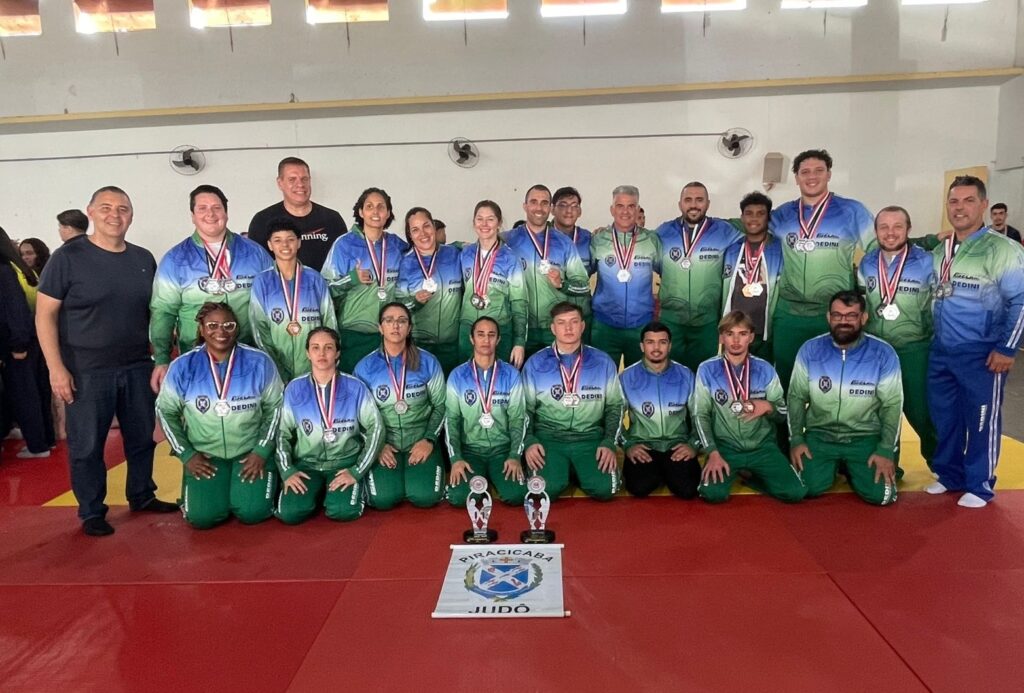 A imagem mostra um grupo de pessoas, provavelmente uma equipe de judô, posando para uma foto após uma competição. Eles estão vestindo jaquetas de judô nas cores verde e azul com detalhes em branco e medalhas ao pescoço. Há troféus e uma faixa com o nome "Piracicaba Judo" na frente do grupo. A maioria das pessoas na foto parece estar orgulhosa e feliz, com alguns membros da equipe sorrindo e outros segurando suas medalhas. A foto foi tirada em um ambiente interno, possivelmente uma quadra de esportes.