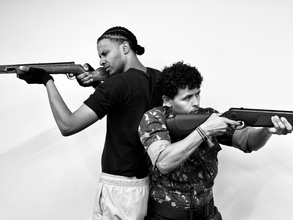 Na imagem aparece dois homens apontando arma de fogo. O primeiro de cabelos amarrado , de camiseta preta e calça clara e o segundo, camisa preta com detalhes em, branco e calça escura. Ambos aponta as armas para cafa um dos lados