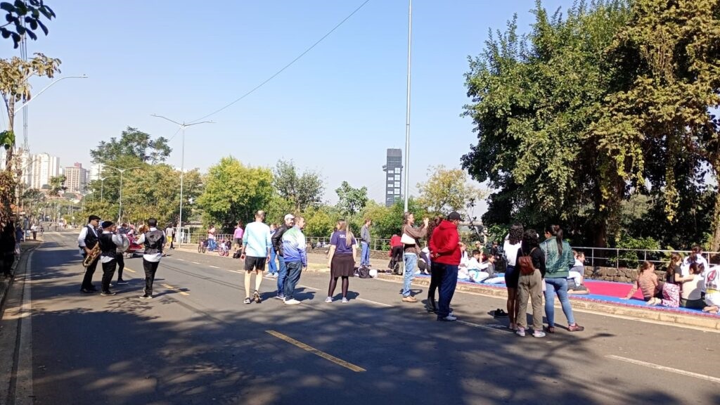 A imagem mostra uma rua pavimentada em um dia ensolarado, com várias pessoas reunidas. Há um grupo de músicos tocando instrumentos de sopro na rua, e um grupo maior de pessoas está sentado ou em pé perto da lateral da rua, algumas em tapetes. Há árvores e vegetação ao longo da rua, e edifícios podem ser vistos ao fundo. O céu está claro e azul. A cena parece ser um evento público ou uma reunião comunitária ao ar livre. Há uma torre alta visível ao fundo, à distância. A atmosfera geral da imagem é tranquila e pacífica. As pessoas parecem estar relaxando e desfrutando do dia ensolarado.