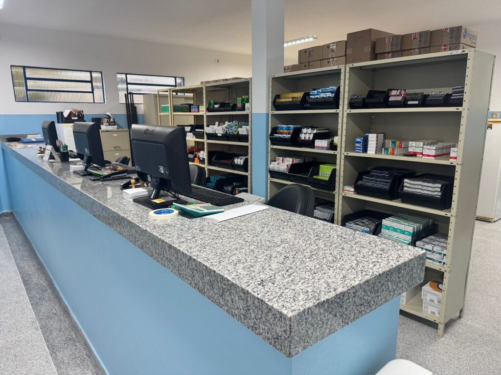 A imagem mostra o interior de um estabelecimento, possivelmente uma farmácia ou drogaria, com um balcão de atendimento e prateleiras de armazenamento. No balcão, há três computadores com monitores LG, teclados e mouses, além de calculadoras, caixas de papel e rolos de fita. O balcão em si é feito de granito com uma base azul clara. Atrás do balcão, há estantes metálicas repletas de caixas de medicamentos e outros produtos, organizados em bandejas pretas. Algumas caixas maiores estão empilhadas no topo das estantes. Há também janelas com venezianas no fundo da sala, permitindo a entrada de luz natural. A iluminação artificial é feita por lâmpadas fluorescentes no teto.