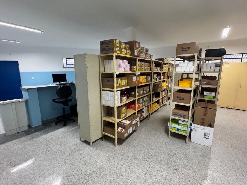 A imagem mostra um escritório ou depósito organizado com várias prateleiras cheias de caixas e produtos. As prateleiras são de metal e estão dispostas em fileira, com uma variedade de caixas de diferentes tamanhos e marcas, como "Hypera" e "Geolab". No lado esquerdo da imagem, há uma bancada com um computador e uma cadeira de escritório, sugerindo que este é um espaço de trabalho onde os produtos armazenados são gerenciados. Há também uma porta azul e uma área de armazenamento branco na extremidade esquerda. O chão é de granilite e o teto possui iluminação fluorescente. A atmosfera geral é de organização e eficiência.