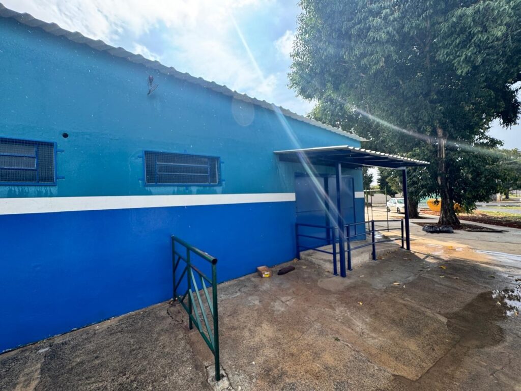 A imagem mostra a fachada de um edifício de cor azul vibrante, com uma faixa branca horizontal dividindo a parte superior e inferior da parede. O edifício possui duas janelas retangulares com grades de proteção, que parecem ser feitas de metal. Há uma marquise metálica que cobre a entrada principal, a qual é composta por duas portas azuis. Um corrimão azul acompanha a rampa de acesso à entrada, e outro corrimão, de cor verde, está posicionado na lateral esquerda da imagem, próximo à parede. O chão à frente do edifício é de concreto, com algumas poças de água visíveis, sugerindo que pode ter chovido recentemente. Ao lado do edifício, à direita, há uma árvore frondosa com um tronco grosso. Mais ao fundo, é possível ver um carro branco, um contêiner amarelo e outras áreas externas, indicando que o local pode ser um espaço público ou uma instalação externa. O céu está parcialmente nublado, com raios de sol atravessando as nuvens.
