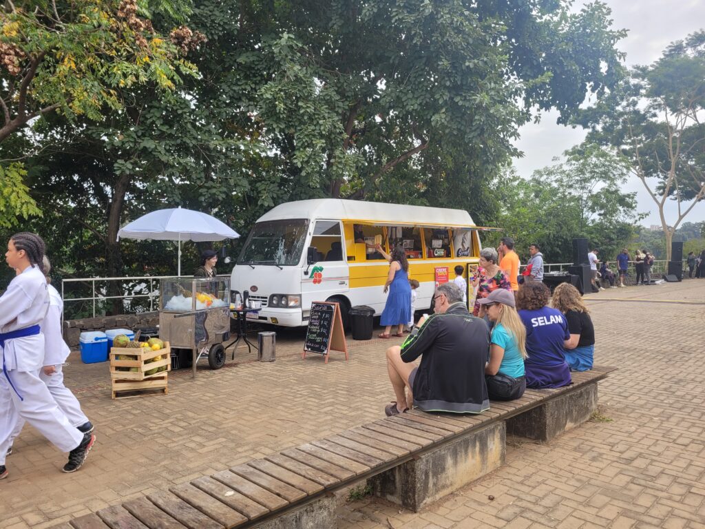 A imagem mostra uma cena ao ar livre em um dia nublado, com um food truck amarelo e branco estacionado sob árvores frondosas. Há pessoas sentadas em um banco de madeira, outras em pé perto do food truck, aparentemente esperando ou comprando comida. Uma pessoa veste um quimono branco e parece estar caminhando. Há também uma barraca menor com frutas e uma pessoa atrás dela. O ambiente parece ser um evento ou feira ao ar livre em um parque ou área semelhante. A atmosfera é casual e relaxada.