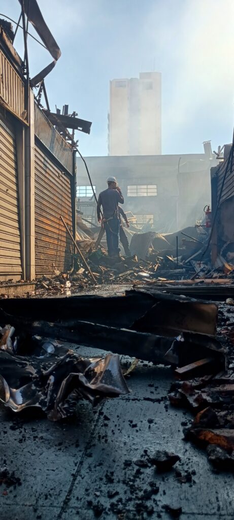 A imagem mostra um cenário pós-incêndio, com um homem vestindo capacete branco e roupas de trabalho em pé no meio dos escombros. Ele parece estar inspecionando o local, com fumaça ainda pairando no ar. A estrutura ao redor está severamente danificada, com metal retorcido e materiais queimados espalhados pelo chão. Ao fundo, um prédio alto é visível, parcialmente obscurecido pela fumaça. A cena geral sugere uma recente catástrofe, com o homem representando a resposta ou avaliação do evento.