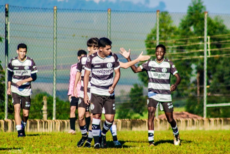 A imagem mostra um grupo de jovens jogadores de futebol, vestindo uniformes listrados em preto e branco com o logo da Raizen. Estão num campo gramado, com uma cerca de arame atrás deles. Um jogador está dando um high-five para outro, enquanto os outros jogadores observam. O fundo mostra árvores e um céu claro. A cena sugere um treino ou jogo informal de futebol juvenil. A iluminação e a qualidade da foto são boas, mostrando detalhes dos uniformes e expressões faciais dos jogadores. A imagem transmite uma atmosfera de energia e camaradagem entre os jovens atletas.