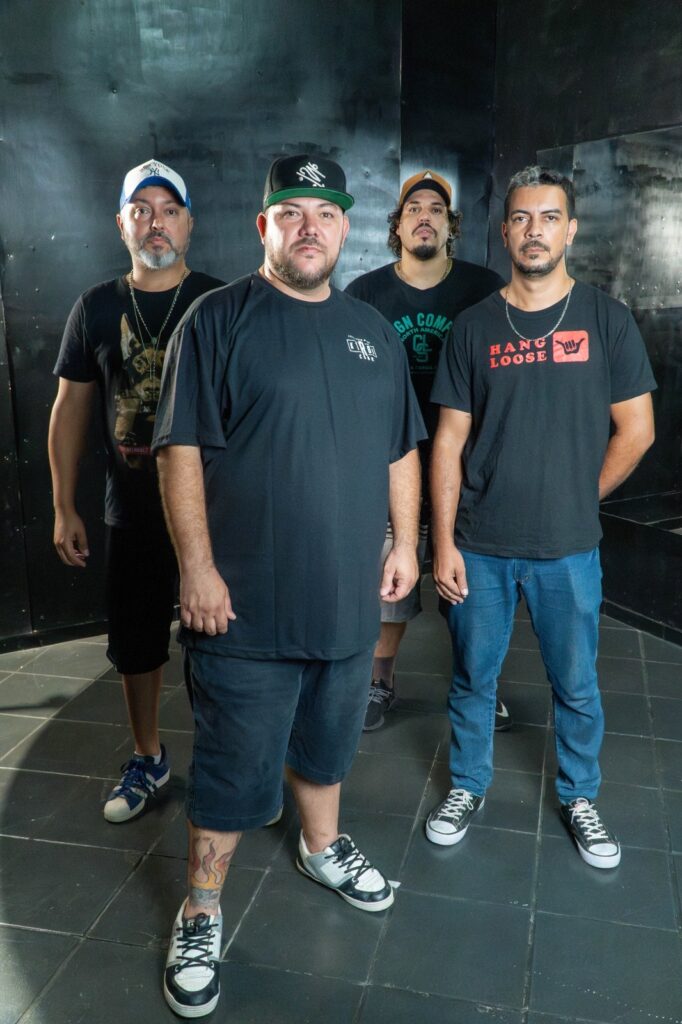 A imagem mostra quatro homens em pé, posicionados lado a lado, voltados para a frente. O fundo é uma parede escura e texturizada, possivelmente de metal. O chão é composto por quadrados escuros, que refletem a luz de forma sutil. O homem mais à esquerda usa um boné branco e azul com o logo da New York Yankees, uma camiseta preta com a imagem de um cachorro e um colar. Ele está com as mãos para baixo, ao lado do corpo. O homem no centro, ligeiramente à frente dos outros, veste um boné preto com um logo verde. Ele usa uma camiseta preta com um pequeno logo branco no peito e shorts escuros. Sua perna esquerda está visível, com uma tatuagem colorida de chamas. Ele está com as mãos cruzadas na frente do corpo. O terceiro homem da esquerda para a direita usa um boné marrom e uma camiseta preta com a inscrição "GN COMA NORTH AMERICA" em verde. Ele está com as mãos atrás das costas.