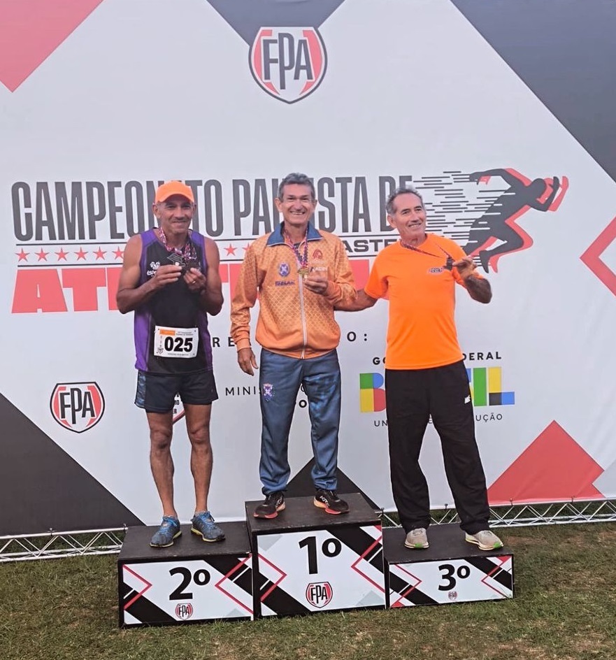 A imagem mostra três homens em um pódio de corrida, aparentemente em uma cerimônia de premiação. Eles estão posicionados em ordem decrescente de classificação, com o vencedor no centro. Todos usam medalhas e roupas esportivas. O homem em primeiro lugar veste uma jaqueta laranja e calças esportivas azuis e cinzas. O segundo colocado usa uma camiseta roxa e shorts pretos. O terceiro colocado veste uma camiseta laranja e calças pretas. O cenário é uma estrutura de banner com o logotipo de uma organização (provavelmente a entidade organizadora da corrida), "FPA", e a inscrição "CAMPEONATO PAULISTA DE ATLETISMO" indicando que se trata de um campeonato de atletismo. O design do banner inclui um desenho estilizado de um corredor. Os números "2º", "1º" e "3º" estão claramente visíveis nos degraus do pódio, indicando as colocações dos corredores. O número "025" está visível na camiseta do corredor que ficou em segundo lugar. O fundo é um gramado. A imagem sugere uma atmosfera de competição esportiva.