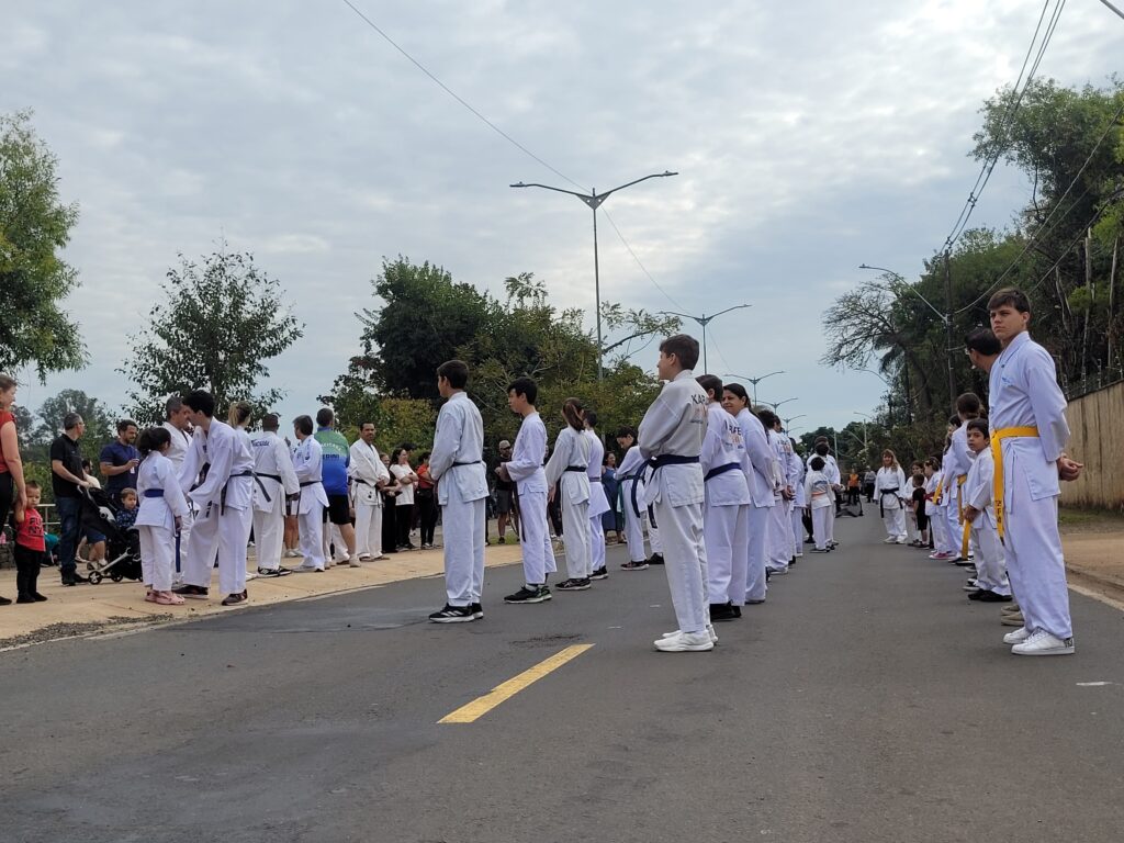 A imagem mostra um grupo de pessoas, principalmente crianças e adolescentes, vestindo uniformes de karatê brancos, alinhados em duas fileiras opostas em uma rua. Eles parecem estar participando de algum tipo de evento ou demonstração. Há também um grupo menor de adultos, alguns vestindo roupas esportivas, observando as crianças. A cena se passa em uma rua arborizada, com um céu parcialmente nublado ao fundo. A atmosfera geral é calma e organizada. Os praticantes de karatê estão em posição de atenção, alguns com faixas coloridas. A imagem sugere uma atividade de treinamento ou um evento público relacionado a artes marciais.