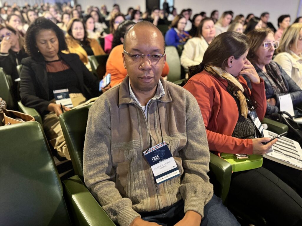 A imagem mostra um homem sentado em uma cadeira em uma sala de conferências lotada. Ele está usando um casaco de lã bege e óculos. Ele está usando um crachá de identificação com o nome "Guilherme D. Manel" e "FNAS São Paulo". O homem está olhando diretamente para a câmera. Ao seu redor, há várias outras pessoas sentadas em cadeiras, a maioria das quais parece estar participando de uma conferência ou apresentação. As mulheres ao seu redor estão vestindo roupas de negócios e algumas estão usando óculos. A atmosfera parece formal e profissional. A iluminação é suave e a imagem é nítida. A imagem parece ter sido tirada em um evento corporativo ou acadêmico.