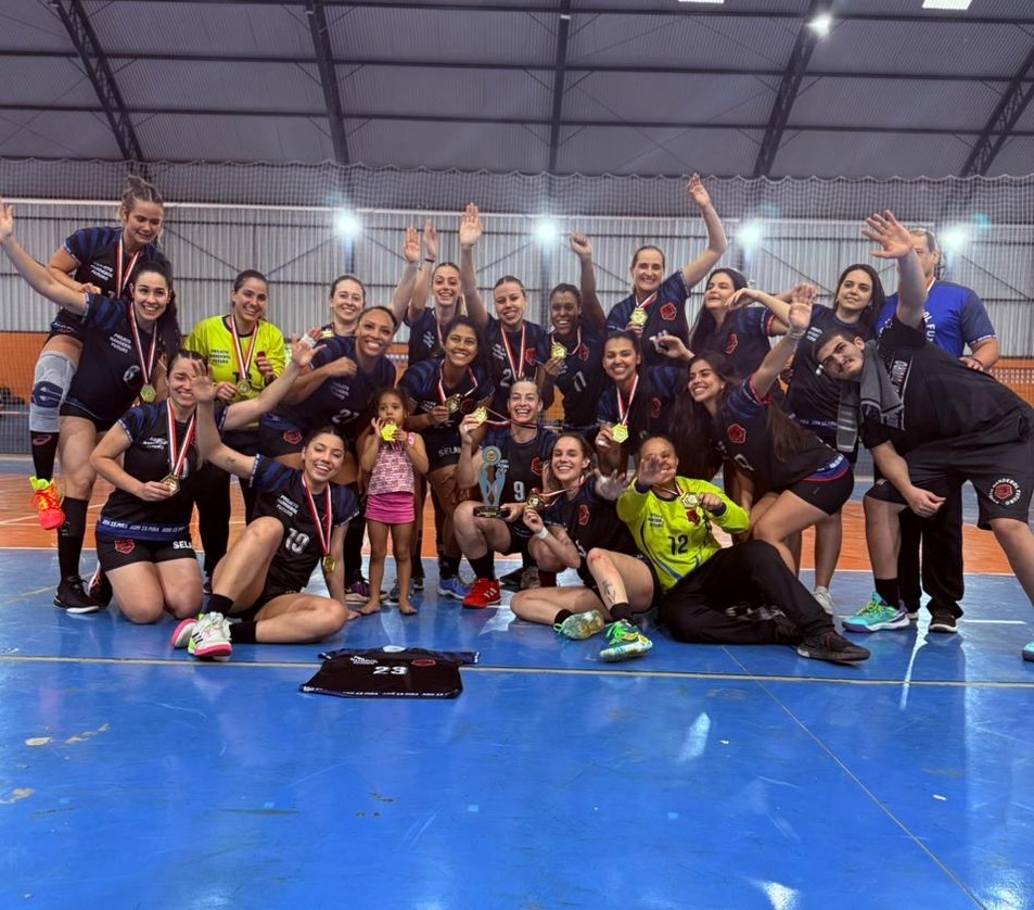 A imagem mostra um grupo de mulheres, aparentemente um time de handebol, comemorando uma vitória. Elas estão reunidas em uma quadra esportiva, muitas com medalhas de ouro ao pescoço e segurando um troféu. Algumas estão sorrindo e abraçando, demonstrando alegria e camaradagem. Há também alguns homens na foto, possivelmente treinadores ou membros da equipe de apoio. Estão todos vestidos com uniformes esportivos, predominantemente pretos e com detalhes em dourado. O fundo é uma quadra esportiva, com paredes e teto visíveis. A atmosfera geral da imagem é de celebração e sucesso. A iluminação é boa, e a foto parece ter sido tirada em um momento espontâneo de comemoração. Há uma camisa de jogo no chão na frente do grupo.