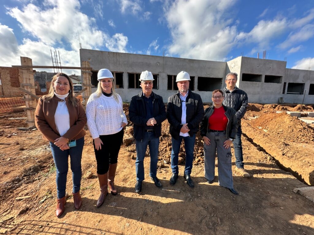 A imagem mostra seis pessoas em frente a uma construção. Cinco delas usam capacetes brancos. As mulheres estão vestidas com roupas sociais, enquanto os homens usam jaquetas. Uma mulher usa uma blusa vermelha e calça jeans. Os homens estão em pé, lado a lado, enquanto as mulheres estão posicionadas em ambos os lados dos homens. A construção ao fundo parece ser uma escola ou prédio público, com paredes de concreto e janelas. O local parece ser uma obra em andamento, com terra e materiais de construção visíveis. O céu está parcialmente nublado, e a luz do sol indica que a foto foi tirada durante o dia. A cena sugere uma visita oficial a um local de construção, possivelmente uma inauguração ou um evento de acompanhamento de obra.