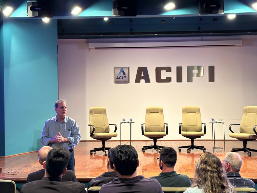 O homem está em pé em um palco, de frente para uma audiência. Ele está vestindo uma camisa xadrez azul e jeans. Suas mãos estão juntas na frente dele. Ele parece estar falando com a audiência. O palco tem um chão de madeira e uma parede branca ao fundo. Há um logotipo "ACPI" na parede, juntamente com as letras "ACPI" em metal prateado. Há quatro cadeiras de escritório giratórias em frente à parede, com mesas laterais entre elas. Há também um projetor e caixas de som no teto. A audiência está sentada em cadeiras em frente ao palco. Eles parecem estar prestando atenção ao homem no palco. A imagem está bem iluminada e o foco está no palestrante.