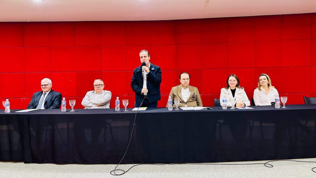 A imagem mostra seis pessoas sentadas em uma mesa longa, com um fundo vermelho texturizado. Um homem está em pé no centro, segurando um microfone e falando. Ele usa uma jaqueta escura sobre uma camisa clara. À sua esquerda, há dois homens. O primeiro, mais próximo do centro, usa um terno escuro com gravata. O segundo, mais à esquerda, veste uma camisa clara e uma jaqueta. Ambos usam óculos. À direita do homem em pé, há duas mulheres e um homem. O homem, mais próximo do centro, usa uma jaqueta xadrez sobre uma camisa branca e um lenço no bolso. As duas mulheres estão sentadas mais à direita. A primeira usa um casaco branco sobre uma blusa escura, e a segunda veste uma camisa branca com um colar. Na frente da mesa, há uma toalha preta. Em cima da mesa, há garrafas de água, copos e papéis. A iluminação é clara, destacando os participantes e o fundo vermelho.