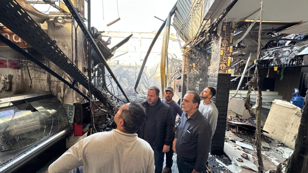 A imagem mostra um grupo de homens observando os estragos causados por um incêndio em um mercado ou centro comercial. O local está severamente danificado, com estruturas metálicas retorcidas, paredes queimadas e destroços espalhados