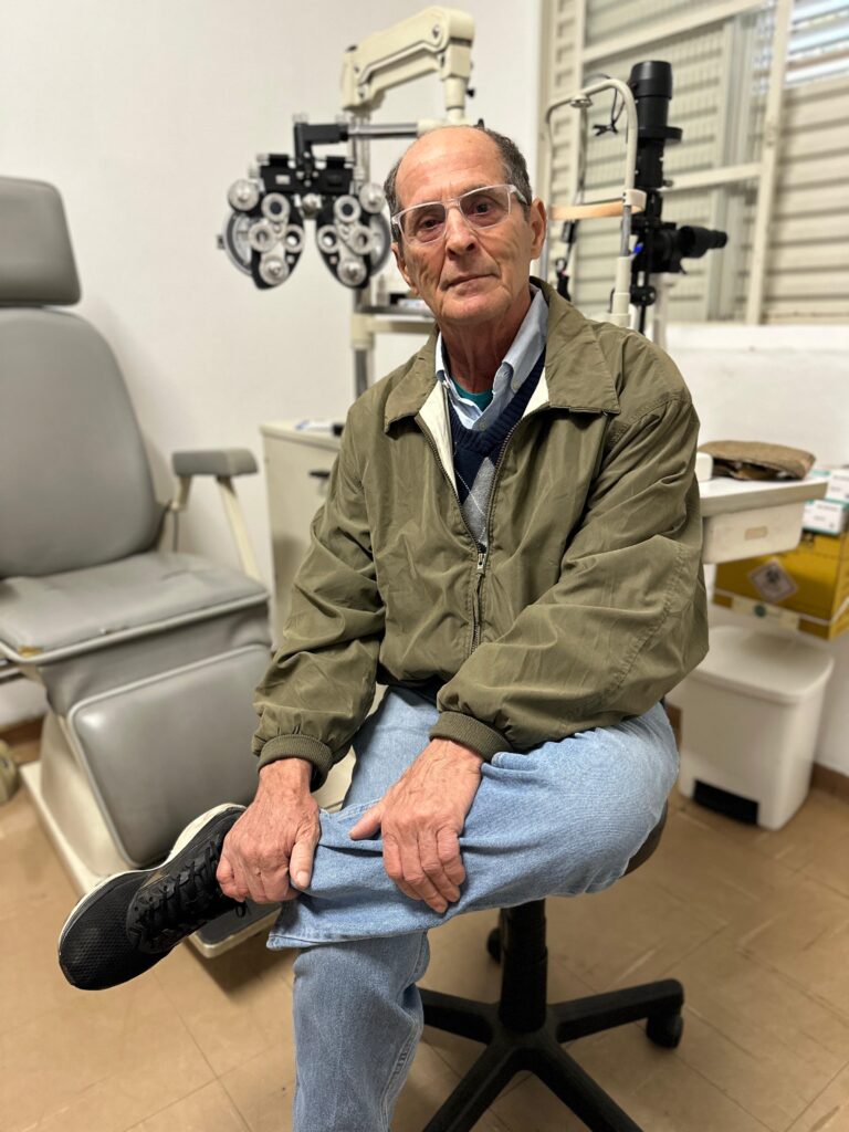 A imagem mostra um homem de meia-idade sentado numa cadeira giratória numa sala de exame oftalmológico. Ele usa óculos, uma jaqueta verde-oliva e calças jeans azuis. A cadeira é de um tom cinzento claro e está posicionada em frente a um aparelho de exame de vista, que é visível ao fundo. O ambiente parece ser um consultório médico, com um ar limpo e organizado. A expressão do homem é séria e ele parece estar à espera ou a ser examinado. Há também uma mesa lateral com alguns objetos, possivelmente materiais médicos. O chão é de azulejo bege claro. A iluminação é suave e difusa, sem sombras fortes. A imagem é nítida e bem focada, com cores realistas.