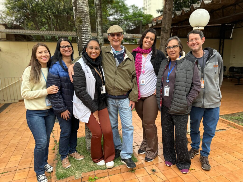 A foto mostra um grupo de sete pessoas sorrindo para a câmera, posicionadas em frente a um muro de tijolos e uma árvore. No centro da imagem está um homem mais velho, de boné e casaco verde-oliva, abraçando duas mulheres jovens de cada lado. As mulheres estão usando roupas casuais e sorrindo. Duas outras mulheres estão em pé atrás delas, ligeiramente à direita, e um homem está em pé atrás delas, ligeiramente à esquerda. Todos parecem estar em um ambiente externo, possivelmente um pátio ou jardim. O homem no centro parece ser o foco principal da foto, com as outras pessoas ao seu redor. A iluminação da foto é natural e suave, indicando que a foto foi tirada ao ar livre em um dia claro. A composição da foto é informal e amigável, capturando um momento de alegria e camaradagem entre as pessoas retratadas. Há um elemento de informalidade na maneira como o grupo está posicionado, com braços ao redor uns dos outros, indicando um relacionamento próximo e amigável. O fundo da foto é um pouco desfocado, mas é possível ver árvores, um prédio e um pouco de vegetação. A foto tem uma atmosfera geral de positividade e amizade.