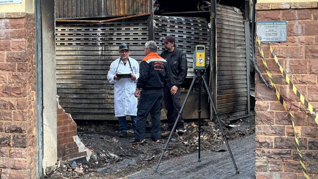 A imagem mostra uma cena de investigação após um incêndio. Três homens estão em frente a uma loja com a porta de metal (rolante) danificada pelo fogo e detritos no chão. Um dos homens, vestindo um jaleco branco, segura um tablet. Outro homem, com uma jaqueta de "Proteção Civil" (Defesa Civil) com a inscrição "PRACACABA-SP", está ao lado dele. O terceiro homem, de boné e jaqueta preta, está atrás deles. Um scanner a laser, montado em um tripé, está posicionado em frente à loja. A fachada da loja é de tijolos vermelhos, e uma fita de isolamento amarela e preta está visível na parede de tijolos à direita. Uma placa com informações sobre o "MERCADO MUNICIPAL DE PIRACICABA" está afixada na parede.
