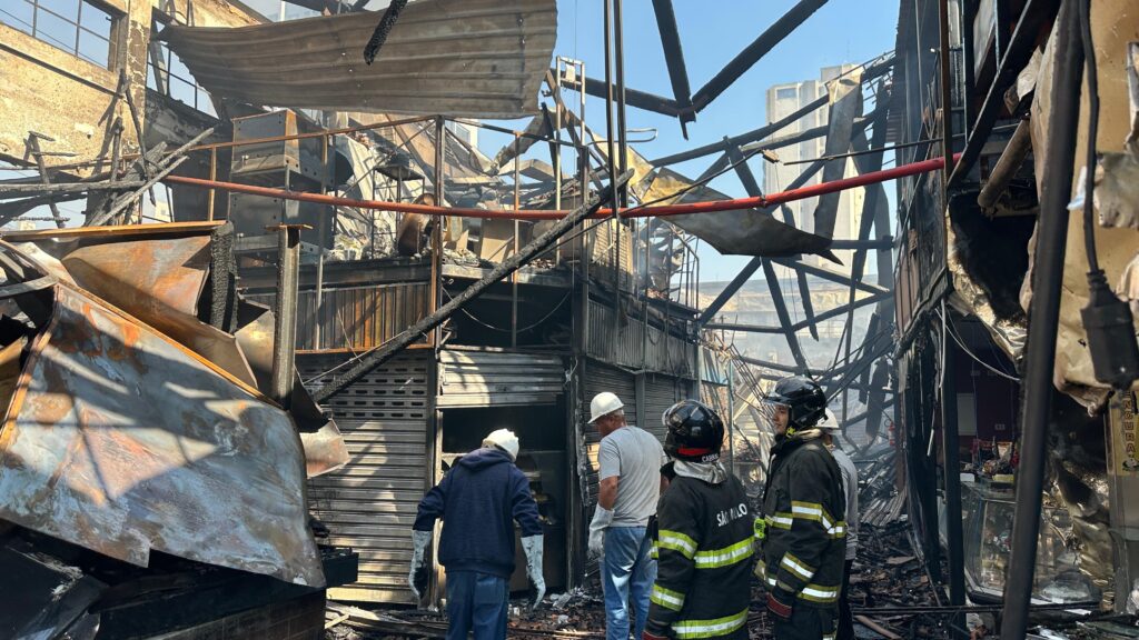 A imagem mostra o interior de um edifício que sofreu um incêndio devastador. A estrutura está em ruínas, com vigas de metal retorcidas e chamuscadas penduradas. O teto desabou em vários lugares, expondo o céu azul e edifícios altos ao fundo. Detritos carbonizados e cinzas cobrem o chão. No primeiro plano, três pessoas vestindo equipamentos de proteção estão examinando os danos. Duas delas parecem ser bombeiros, com capacetes pretos e jaquetas de proteção com listras refletivas. Uma delas tem "SÃO PAULO" escrito nas costas. A terceira pessoa usa um capacete branco e parece ser um trabalhador da construção ou um inspetor. Todos parecem estar avaliando a extensão da destruição. A cena transmite uma sensação de desolação e destruição, com os restos do que foi um edifício agora reduzidos a uma carcaça carbonizada.