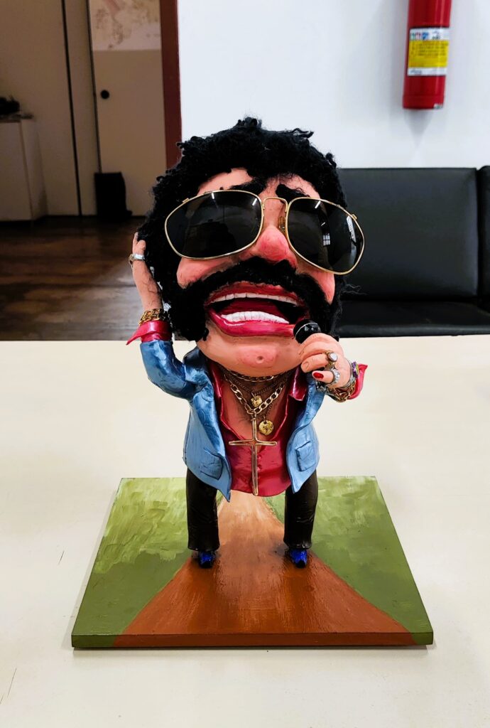 A imagem mostra uma escultura de um homem com óculos escuros, bigode e uma cruz dourada. Ele está vestindo uma jaqueta azul e uma camisa vermelha. Ele está segurando um microfone e parece estar cantando. A escultura está em cima de uma base quadrada com uma pintura de uma estrada. O fundo é uma parede branca com uma porta de madeira e um extintor de incêndio. Há também um sofá preto na imagem. A escultura é feita de um material que parece argila ou massa de modelar. Os detalhes são bem feitos e a escultura é muito expressiva. Parece ser uma obra de arte única e criativa.