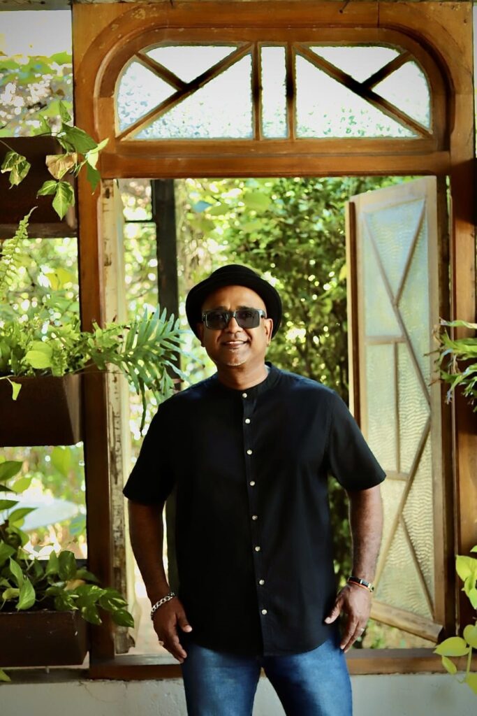 A imagem mostra um homem sorrindo, vestindo um chapéu preto, óculos escuros, uma camisa preta de botões e calças jeans azuis. Ele está em pé em frente a uma janela com moldura de madeira e vidro texturizado, com plantas verdes em vasos ao redor. A luz do sol ilumina a cena, criando um ambiente natural e relaxante. As mãos do homem estão descansando nos quadris, e ele usa uma pulseira prateada em seu pulso esquerdo.