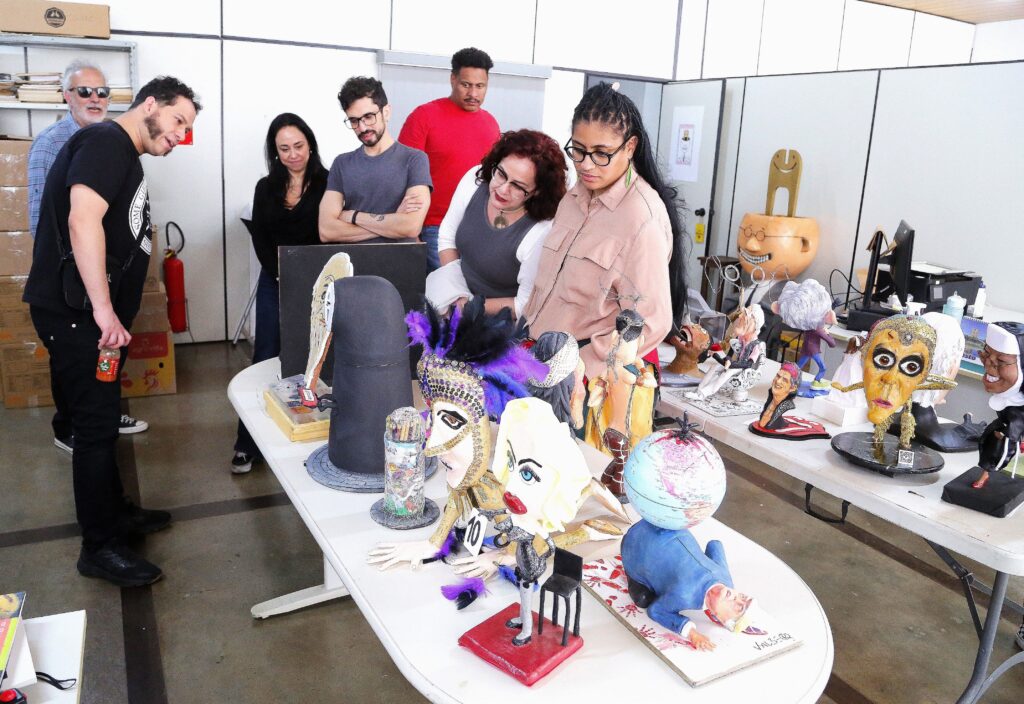 A imagem mostra um grupo de pessoas observando uma coleção de bonecos e esculturas em uma feira ou exposição. Os bonecos são diversos, com alguns apresentando características exageradas e cômicas, como cabeças grandes e expressões faciais marcantes. Há também objetos que parecem ser chapéus decorados com penas e outros enfeites. As pessoas na imagem parecem estar engajadas na observação, com alguns apontando e conversando sobre as peças expostas. O ambiente parece ser um espaço interno, possivelmente uma galeria ou um salão de eventos, com mesas brancas exibindo os trabalhos e caixas e extintores de incêndio ao fundo.