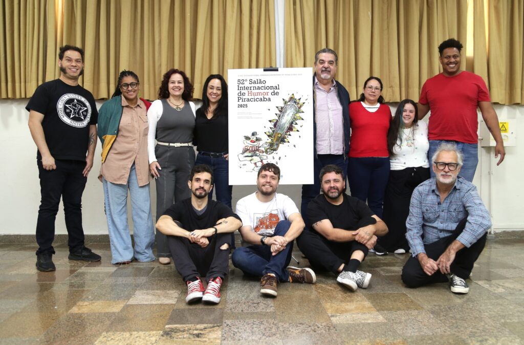 A imagem mostra um grupo de 11 pessoas posando para uma foto em frente a um banner que anuncia o "52º Salão Internacional de Humor de Piracicaba 2025". As pessoas estão em duas fileiras: seis em pé atrás e cinco sentadas na frente. Na fileira de trás, da esquerda para a direita, estão: um homem com cabelo cacheado e espetado, vestindo uma camiseta preta com um logo circular; uma mulher com óculos e cabelo trançado, vestindo uma camisa de botões marrom; uma mulher com cabelo cacheado e escuro, vestindo uma blusa cinza e calça cinza; uma mulher com cabelo escuro e liso, vestindo uma blusa preta e jeans; um homem com cabelo grisalho e curto, vestindo uma camisa social clara e jaqueta azul; e um homem com cabelo curto e escuro, vestindo uma camiseta vermelha e jeans. Na fileira da frente, da esquerda para a direita, estão: um homem com barba e cabelo escuro, vestindo uma camiseta preta rasgada no joelho e tênis vermelhos; um homem sorrindo, vestindo uma camiseta branca com estampa gráfica e jeans; um homem com barba e cabelo escuro, vestindo uma camiseta preta e tênis com estampa; e um homem com cabelo grisalho e ondulado, vestindo uma camisa xadrez azul e calça jeans. O banner ao centro exibe uma ilustração de um boneco de neve com cachecol e um pedaço de madeira com uma corrente de motosserra, com o título do evento em destaque. O fundo é composto por cortinas bege e o chão é coberto por um piso xadrez em tons de marrom e bege. Há também um sinal de alerta amarelo visível no canto superior direito.