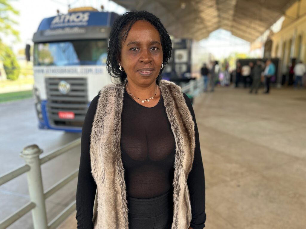 A imagem mostra uma mulher negra de meia-idade, com cabelo curto e escuro, vestindo uma blusa preta transparente por baixo de um colete de pele sintética bege. Ela está em pé em um local que parece ser uma estação rodoviária ou um ponto de ônibus, com um ônibus da empresa Athos ao fundo, ligeiramente fora de foco. O fundo mostra outras pessoas, também ligeiramente desfocadas, sugerindo um ambiente público movimentado. A expressão da mulher é neutra, seu olhar direto para a câmera. A iluminação é natural, e a imagem tem uma qualidade fotográfica casual. A mulher usa brincos pequenos e um colar delicado.