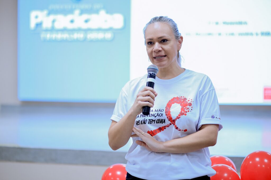 A imagem mostra uma mulher de meia idade, de cabelos grisalhos e curtos, vestindo uma camiseta branca com uma estampa vermelha em formato de laço e a frase "Junte as mãos à prevenção! AIDS não tem cura". Ela segura um microfone e parece estar dando uma palestra ou apresentação. Ao fundo, há uma tela com o logotipo de uma empresa, possivelmente o local do evento. Há balões vermelhos no chão, sugerindo um evento relacionado à conscientização sobre alguma causa, provavelmente o HIV/AIDS, considerando a estampa da camiseta. A expressão da mulher é séria e concentrada. A iluminação é suave, e o ambiente parece ser interno e formal.