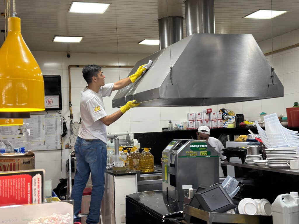 Na imagem, dois homens estão em uma cozinha comercial. Um homem com uma camiseta branca e jeans está em pé em uma escada, limpando uma grande coifa de metal com um pano e luvas amarelas. O outro homem, usando uma camiseta branca e um boné branco, está mais atrás, olhando para uma máquina de moer cana-de-açúcar. A cozinha parece estar em funcionamento, com vários suprimentos visíveis, incluindo garrafas de óleo, pratos empilhados e embalagens de produtos. Há também um cardápio em um suporte na frente esquerda da imagem.