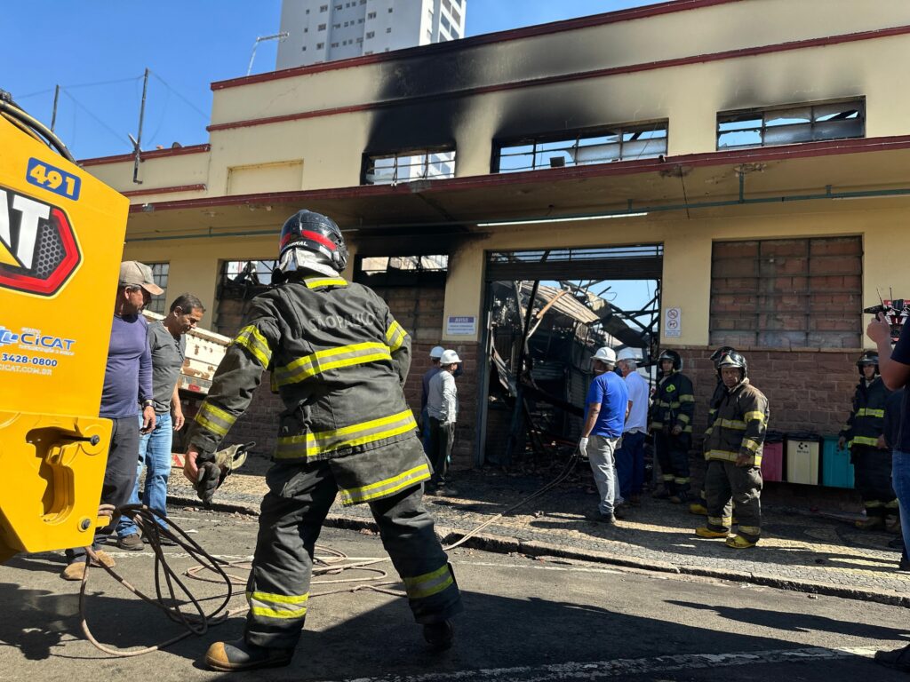 A imagem mostra uma cena após um incêndio em um edifício. Um bombeiro em primeiro plano, vestindo equipamento de proteção completo, incluindo capacete e vestes antichama com faixas refletivas, parece estar manuseando uma mangueira ou cabo. Ele está de costas para o observador, voltado para a entrada do edifício. O edifício em si exibe sinais claros de danos causados pelo fogo, com uma grande área de fuligem preta cobrindo a parte superior da fachada, especialmente acima das janelas. As janelas aparentam estar quebradas ou danificadas. A entrada principal do edifício está aberta, revelando o interior destruído e colapsado, com escombros visíveis. Ao redor da entrada e em frente ao edifício, há um grupo de pessoas. Vários bombeiros com equipamentos semelhantes estão presentes, alguns observando a cena e outros possivelmente em trabalho. Há também civis, incluindo um homem com boné e outro em camiseta cinza, que parecem estar inspecionando os danos. À esquerda da imagem, uma parte de um veículo amarelo, possivelmente um caminhão de bombeiros ou equipamento de apoio, é visível. O veículo tem a inscrição "CICAT" e um número de telefone. O céu está azul e claro, indicando um dia ensolarado. A cena geral sugere uma investigação ou avaliação pós-incêndio.