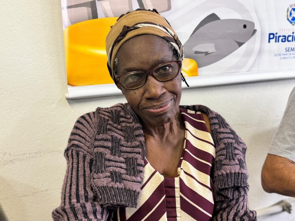 A imagem mostra uma mulher idosa, de pele negra, sorrindo para a câmera. Ela está usando um lenço na cabeça, óculos e um suéter com um padrão geométrico em tons de roxo e preto. Por baixo do suéter, ela usa uma blusa listrada em tons de vinho e creme. Ao fundo, há uma parede branca com um cartaz que exibe um peixe cinza estilizado e o texto "PREFEITURA PIRACICABA".