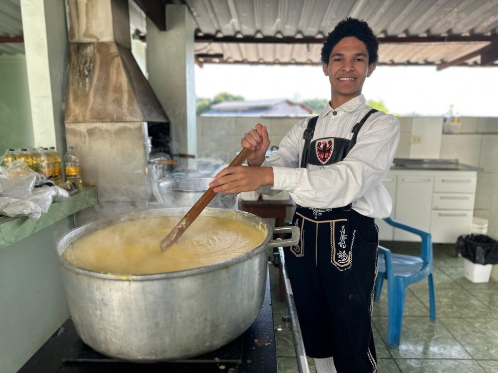 A imagem mostra um jovem sorrindo enquanto mexe uma grande panela de polenta com uma colher de pau. Ele está vestindo uma camisa branca e calças pretas com detalhes bordados, que parecem ser um traje tradicional. A panela está em um fogão ao ar livre, sob uma estrutura coberta. Há outros utensílios de cozinha visíveis ao fundo, incluindo uma chaminé e armários. O ambiente parece ser uma área externa ou pátio de uma casa ou comunidade. A cena sugere a preparação de uma refeição comunitária ou evento. O foco principal da imagem é o jovem e sua atividade de cozinhar, criando uma sensação de calor e comunidade. A iluminação é natural e suave.