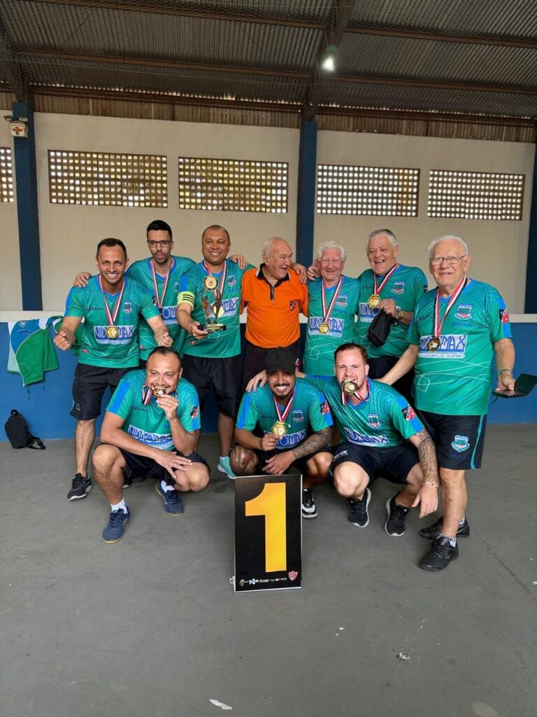 A imagem mostra um grupo de homens comemorando uma vitória em um evento esportivo. Eles estão vestindo uniformes verdes e azuis com o logo "NIDX MAX". Todos usam medalhas de ouro no pescoço, indicando que ganharam o primeiro lugar. Um homem no centro está segurando um troféu, enquanto os outros posam para a foto com sorrisos e expressões de alegria. Há também um grande número "1" em um suporte, reforçando a ideia de que eles são os campeões. O fundo sugere que o evento aconteceu em uma quadra ou ginásio.
