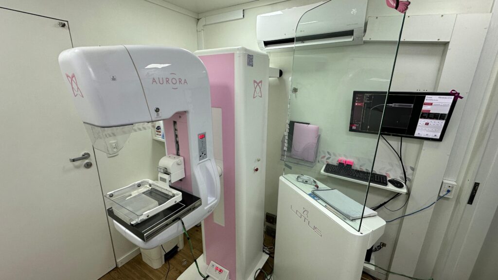 A imagem mostra uma sala de espera pequena e moderna, possivelmente dentro de um trailer ou unidade móvel. As paredes são brancas, com uma parede de destaque em rosa com o texto "Mulheres de Peito". O chão é de madeira escura. Há várias cadeiras pretas de plástico alinhadas ao longo das paredes. Uma recepção branca com dois computadores e uma impressora fica no centro da sala. Há também uma televisão de tela plana montada na parede. A decoração inclui vários quadros rosa com imagens de mulheres e mensagens de autocuidado. A iluminação é brilhante e artificial. A sala é limpa e organizada, dando a impressão de um ambiente profissional e acolhedor.