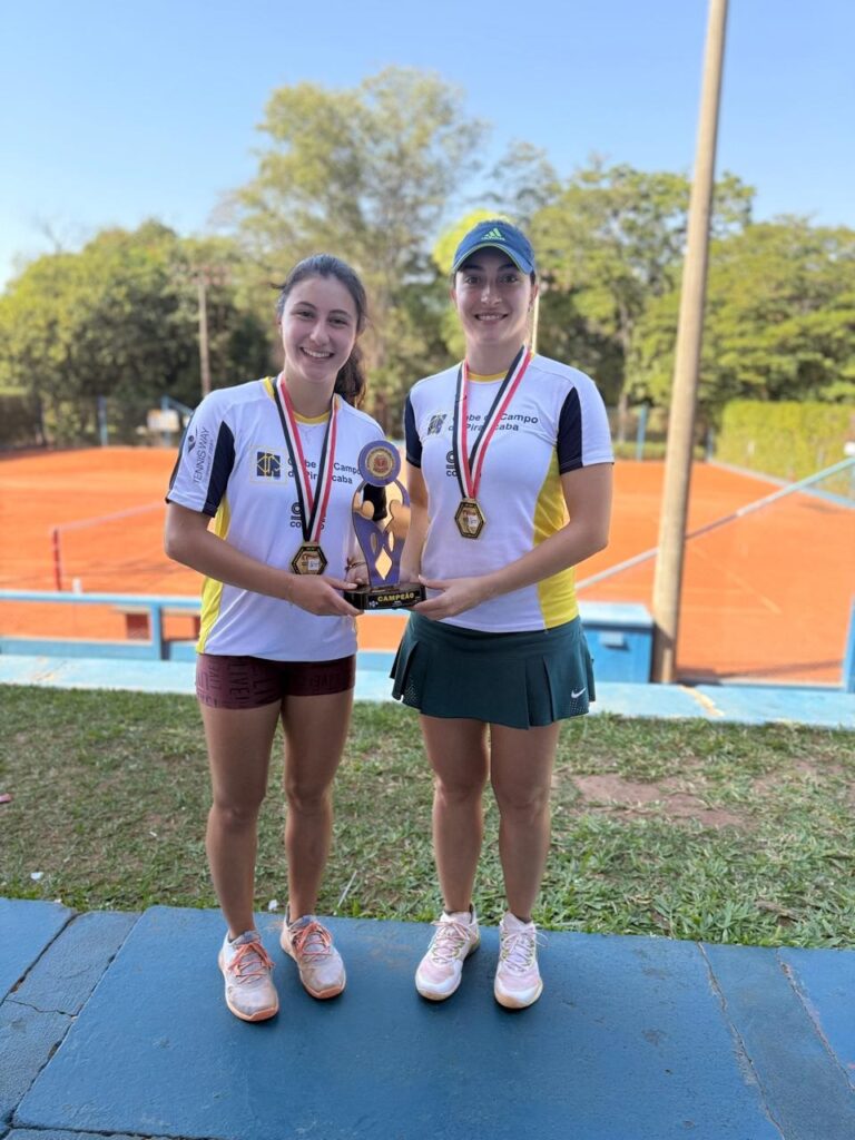 Duas jovens tenistas posam para uma foto, ambas com medalhas de ouro no pescoço e segurando troféus. Elas estão em uma quadra de tênis, com uma quadra de terra batida visível ao fundo, cercada por árvores sob um céu azul claro. As duas usam uniformes de tênis, com a da esquerda ostentando uma medalha e um troféu menor, enquanto a da direita exibe uma medalha e um troféu maior. Ambas estão sorrindo para a câmera, irradiando orgulho e realização.