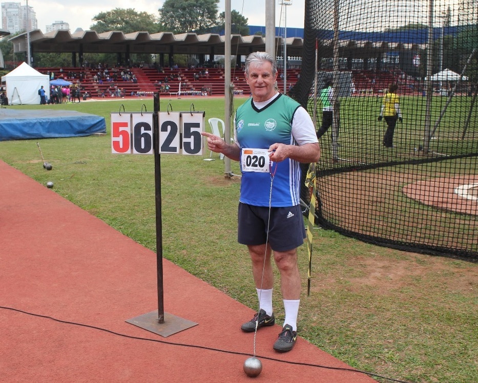 A imagem mostra um homem de meia-idade, de cabelos grisalhos e pele clara, vestindo uma camiseta azul e shorts escuros da Adidas, segurando um peso de lançamento de martelo. Ele está em uma pista de atletismo, com uma rede de proteção atrás dele. Um placar eletrônico mostra o número 5625. Há outros atletas e espectadores ao fundo, sugerindo um evento esportivo de atletismo. O homem parece orgulhoso e satisfeito, possivelmente após um lançamento bem-sucedido. O ambiente é um estádio ao ar livre, com grama e uma pista de corrida vermelha. O dia parece ensolarado. A imagem sugere uma competição de lançamento de martelo, com o homem como o principal foco.