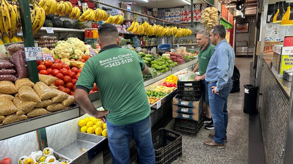 Uma cena vibrante se desenrola em uma banca de mercado movimentada, exibindo uma colorida variedade de produtos frescos. Em primeiro plano, um homem com uma camiseta verde com "Banca do Cláudio" impressa nas costas é visto de costas, com a mão apoiada no quadril. À sua direita, dois outros homens estão em pé, um com uma camisa verde e o outro com uma camisa jeans azul, ambos parecendo estar envolvidos em conversa ou trabalho. A banca está carregada com uma variedade de frutas e vegetais. Cachos de bananas amarelas pendem acima, enquanto abaixo, sacos de pipoca e tomates vermelhos suculentos estão organizados. Couves-flor, pepinos, pimentões, milho e limões adicionam à rica paleta de produtos. Ao fundo, prateleiras estão abastecidas com mais mercadorias, incluindo potes de itens conservados e rolos do que parecem ser produtos de papel. A impressão geral é de abundância e comércio animado.