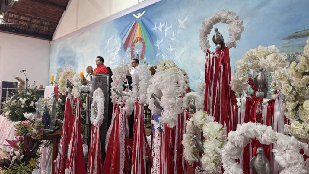A imagem mostra o interior de uma igreja, possivelmente durante uma cerimônia religiosa. No altar, há vários padres e pessoas vestindo roupas formais. Há um grande mural na parede atrás deles, representando pombas brancas em voo e um arco-íris. Na frente do altar, há muitas hastes decoradas com coroas de flores brancas e pombas metálicas. As hastes são adornadas com fitas vermelhas e brancas, criando um efeito visual vibrante e festivo. As flores parecem ser artificiais, e as pombas são de um material metálico prateado. A cena como um todo sugere uma celebração religiosa significativa, possivelmente um evento especial ou festivo dentro da comunidade.