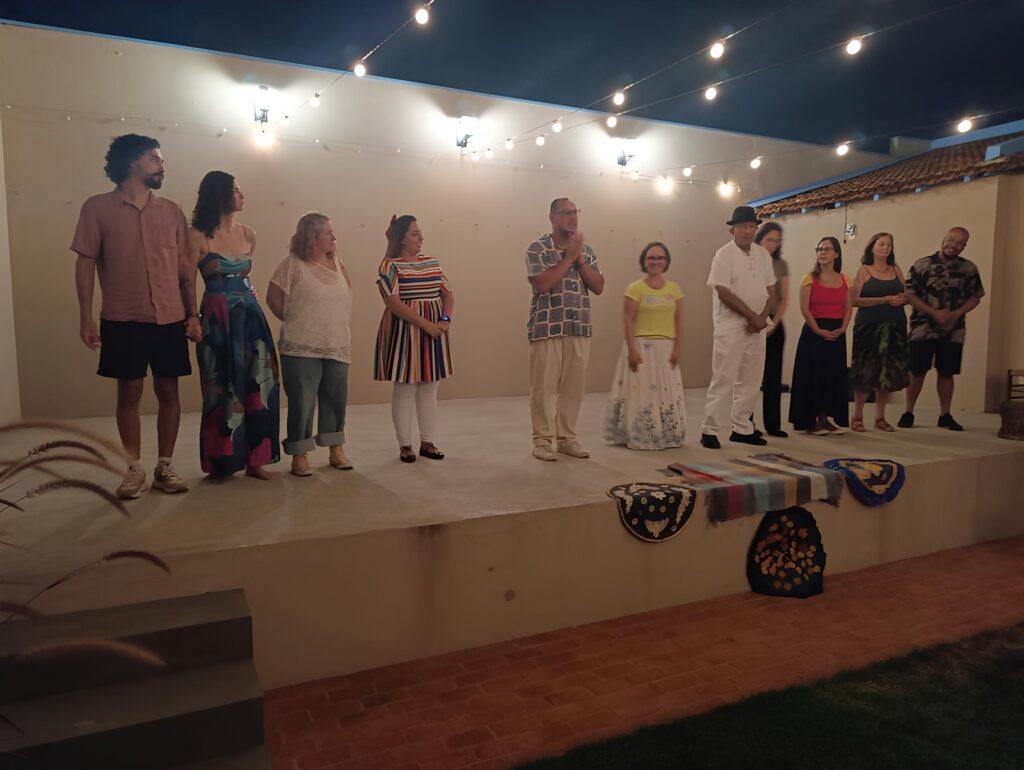 A imagem mostra um grupo de dez pessoas em pé, em uma plataforma elevada, sob a luz de várias lâmpadas penduradas. O fundo é uma parede clara, e acima dela, o céu noturno escuro. À esquerda, um homem com cabelo encaracolado e barba veste uma camisa de manga curta bege e shorts pretos. Ao lado dele, uma mulher com cabelo escuro e encaracolado usa um vestido longo com um padrão abstrato em tons de azul, rosa e verde. Em seguida, uma mulher com cabelo loiro e liso está vestida com uma blusa branca de renda e calças jeans dobradas. Uma mulher com cabelo escuro e um laço vermelho na cabeça usa um vestido listrado vibrante em várias cores. No centro da plataforma, um homem com óculos veste uma camisa xadrez em tons de azul e branco e calças bege. Ao seu lado, uma mulher com cabelo escuro e curto usa uma blusa amarela e uma saia longa branca com um padrão azul. Um homem com um chapéu preto está vestido com uma camisa branca e calças brancas. Uma mulher com cabelo escuro e óculos usa um top vermelho e uma saia longa preta. Outra mulher com cabelo escuro e liso veste um top verde escuro e uma saia estampada. Finalmente, à direita, um homem com cabelo curto e escuro usa uma camisa estampada e shorts pretos. Na frente da plataforma, há alguns objetos decorativos, incluindo tapetes coloridos e um arranjo escuro com detalhes em laranja. O chão em primeiro plano é pavimentado com tijolos de cor terrosa.