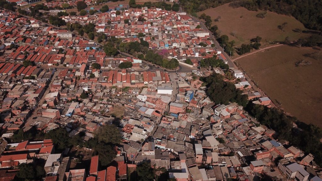 A imagem mostra uma vista aérea de uma favela, com muitas casas pequenas e próximas umas das outras. As casas são construídas com materiais simples, como tijolos e telhas, e muitas delas parecem estar em estado de degradação. Há pouca vegetação na área, e as ruas são estreitas e irregulares. Ao lado da favela, há uma área de campo aberto. A imagem sugere pobreza e falta de infraestrutura na área. A densidade populacional é alta, e as casas estão compactadas. A imagem também mostra uma mistura de casas de diferentes tamanhos e estilos arquitetônicos, indicando uma construção orgânica e informal ao longo do tempo. A falta de planejamento urbano é evidente.
