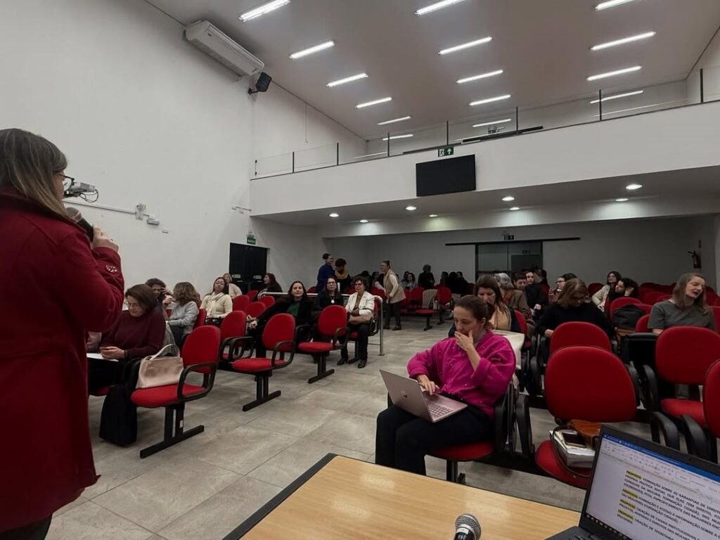 Na imagem aparece um grupo de mulheres sentadas em cadeiras vermelhas, em duas fileiras. A frente aparece uma mulher de óculos de roupa vermelha, com microfone na mão direita, falando ao público, que está acomodado em um espaço que parece um auditório.