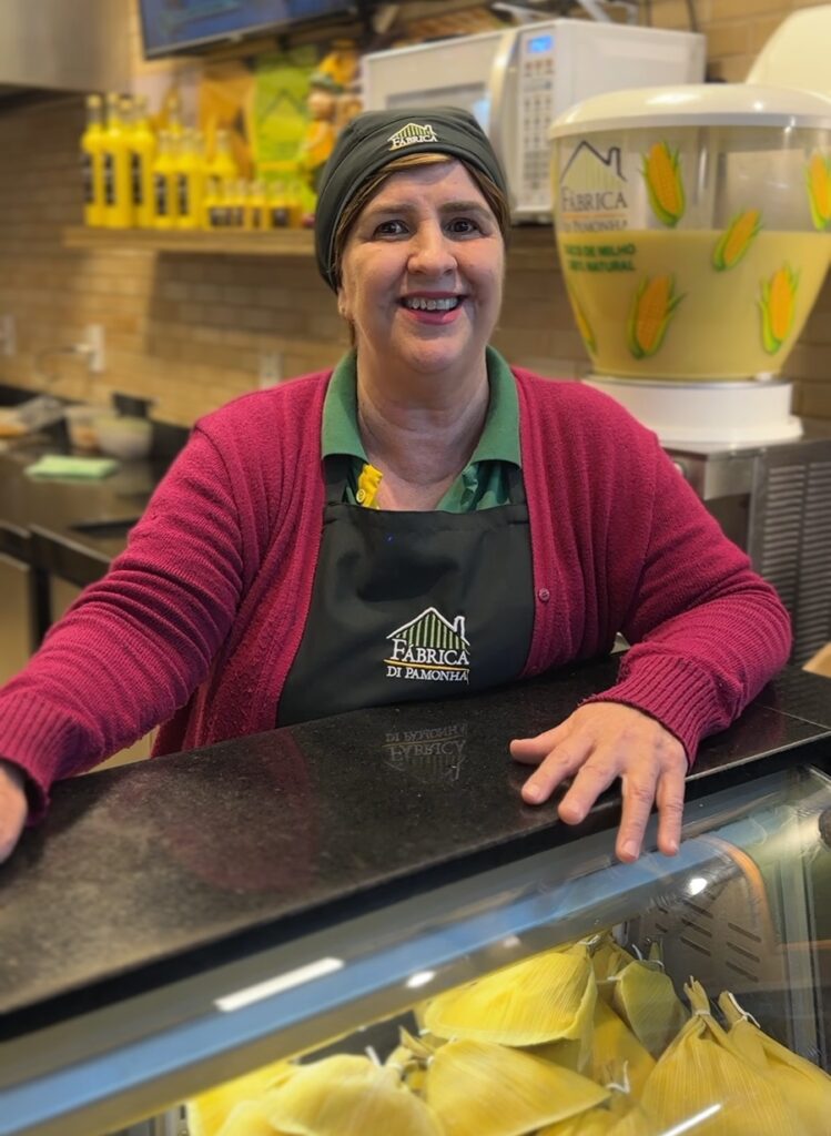 A mulher, com um sorriso radiante, está atrás de um balcão de mármore escuro, vestindo um avental preto com o logotipo "Fábrica di Pamonha" e um cardigã vinho sobre uma camisa verde. Ela usa uma touca preta com o mesmo logotipo bordado. Atrás dela, em uma prateleira, há várias garrafas de um líquido amarelo, possivelmente suco ou bebida, e um dispensador de plástico transparente com milho e a inscrição "Fábrica di Pamonha - Suco de Milho 100% Natural". Na frente do balcão, em uma vitrine refrigerada, há uma grande quantidade de pamonhas embrulhadas em folhas de milho.