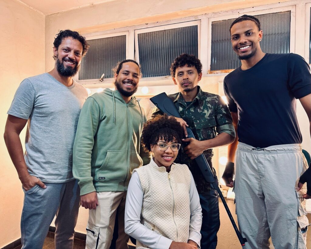 A imagem mostra cinco pessoas em um ambiente que parece ser um camarim ou um local de bastidores. Todas as cinco pessoas estão sorrindo e olhando para a câmera. Da esquerda para a direita: 1. Um homem com cabelo encaracolado e barba, vestindo uma camiseta cinza e calças de moletom cinza. Ele está com a mão no bolso. 2. Um homem com cabelo preso em um rabo de cavalo, vestindo um moletom com capuz verde e calças bege. Ele está sorrindo amplamente. 3. Uma mulher com cabelo afro e óculos, vestindo um colete acolchoado bege sobre uma blusa branca. Ela está sentada em primeiro plano, com as mãos juntas no colo. 4. Um homem vestindo uma uniforme militar camuflado, segurando uma arma de forma relaxada. Ele tem cabelo encaracolado e um bigode. 5. Um homem com cabelo curto e trançado, vestindo uma camiseta preta e calças cargo bege. Ele está sorrindo e tem um piercing na orelha.