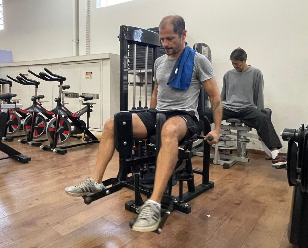 A imagem mostra uma academia com dois homens exercitando-se. Um homem está sentado em uma máquina de extensão de pernas, com os joelhos flexionados e os pés apoiados nos pedais. Ele está usando tênis, shorts e uma camiseta cinza. Um pano azul está pendurado em seu pescoço. O outro homem está sentado em uma máquina de exercícios diferente, parecendo um aparelho de abdução e adução de pernas. Ele está usando calças e uma camiseta cinza. Ambos os homens parecem estar focados em seus exercícios. No fundo, há várias bicicletas ergométricas alinhadas. O piso da academia é de madeira. A iluminação é clara e natural. A atmosfera é calma e tranquila.