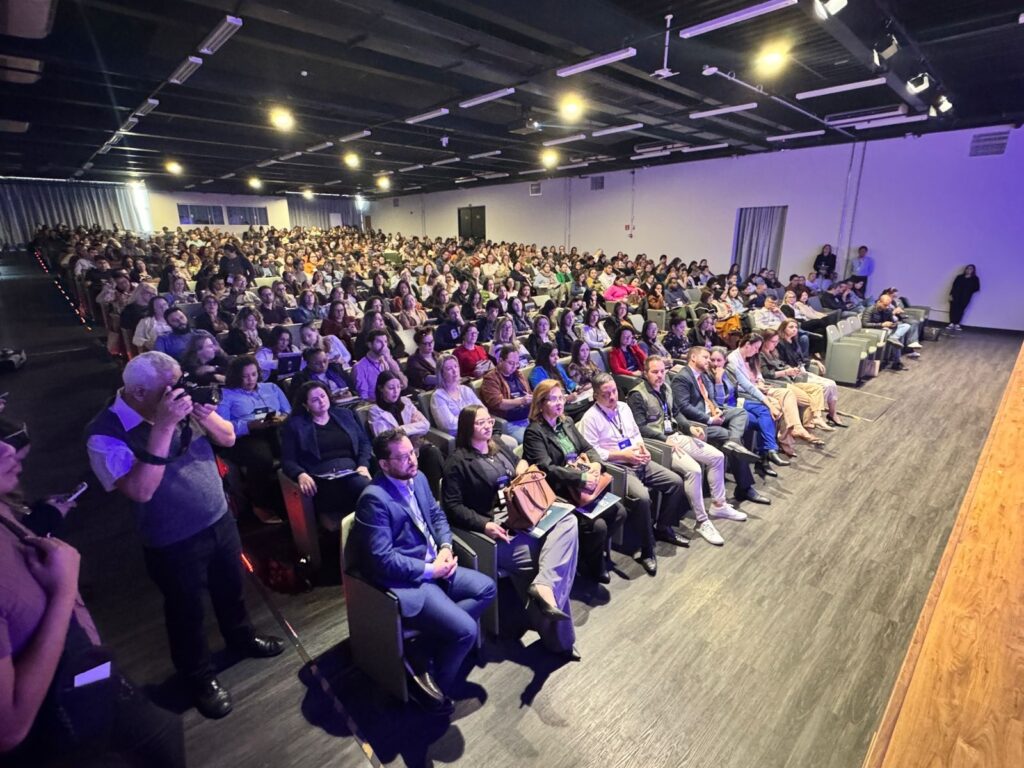 A imagem mostra uma grande plateia de pessoas sentadas em um auditório. A maioria das pessoas está vestida de forma casual, e há uma mistura de homens e mulheres de diferentes idades. As pessoas estão sentadas em fileiras, e há um palco vazio no fundo. Há um fotógrafo tirando fotos da plateia. A iluminação é suave, e a atmosfera é calma e tranquila. A imagem é nítida e bem iluminada, e as cores são vibrantes.